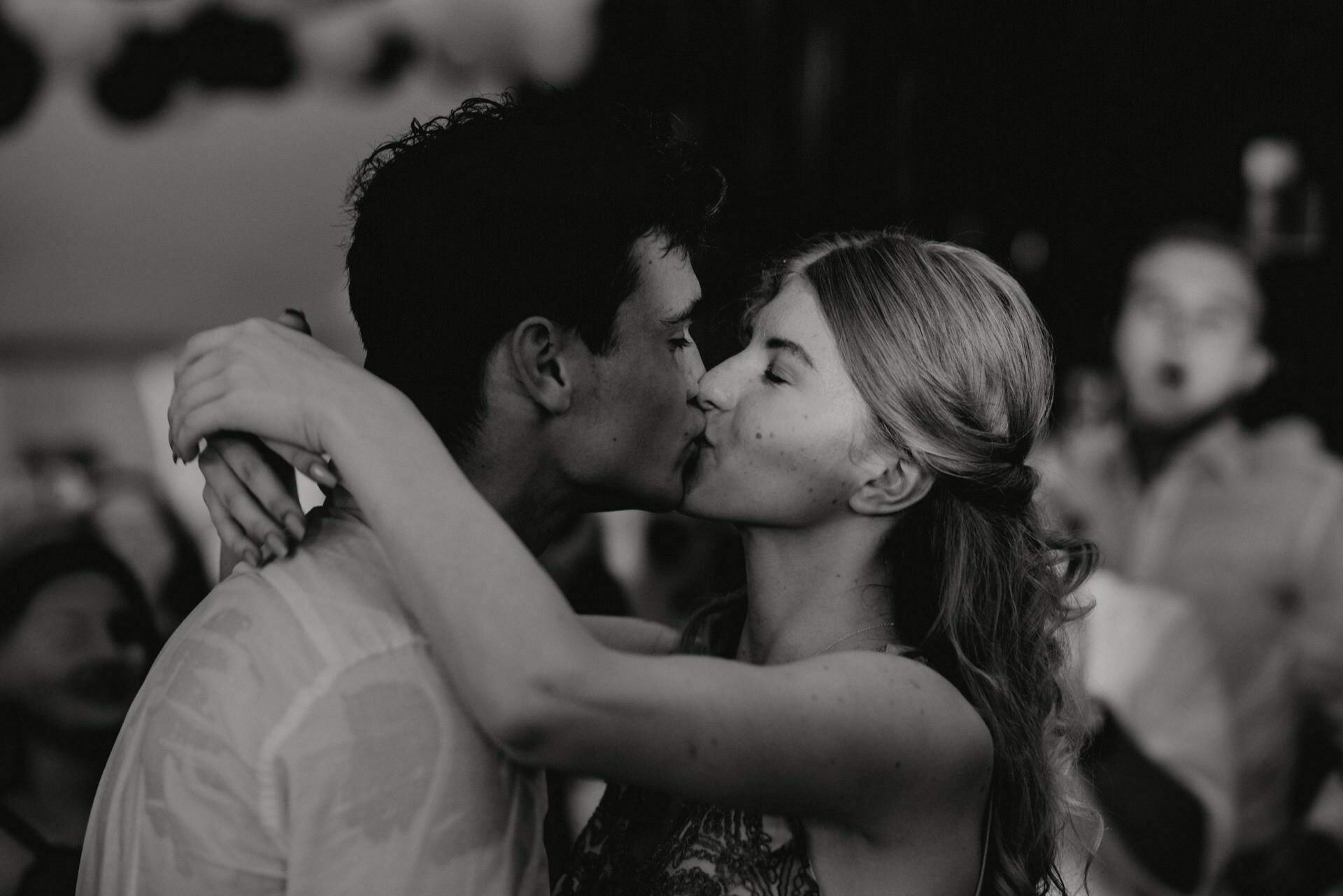 Black and white photo showing a young couple kissing, taken by an event photographer Warsaw. The woman has long hair and her arms are wrapped around the man's neck, while he holds her close. The blurred background with blurred figures and lights adds to the intimate, romantic atmosphere.  