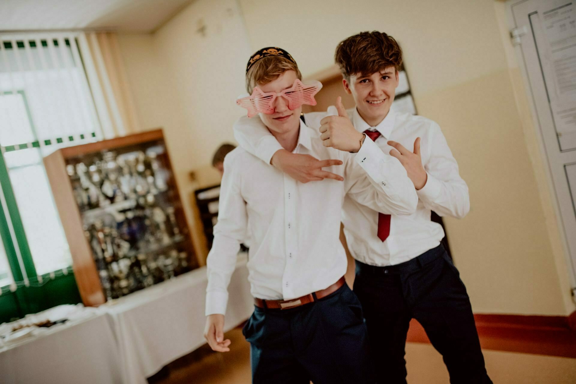 Two young men dressed in white shirts and dark pants pose together in a room. One is putting his arm around the other's shoulder, wearing pink sunglasses, and both are giving a thumbs up. This event photography beautifully captures their mood of celebration, perfect for any photo event.  