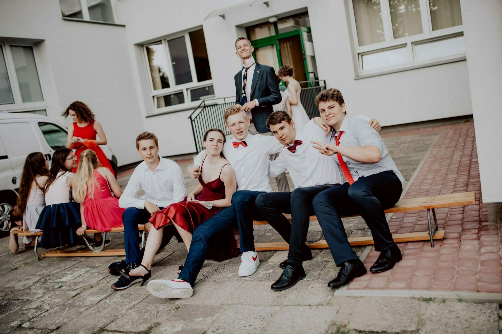 A group of young people are gathered outside the building, sitting on a wooden bench and the ground. Four of them are sitting on the bench, one is standing behind them, and several others are sitting on the ground in the background. They all seem relaxed and happy - the perfect event photo shoot for any event photographer Warsaw.  