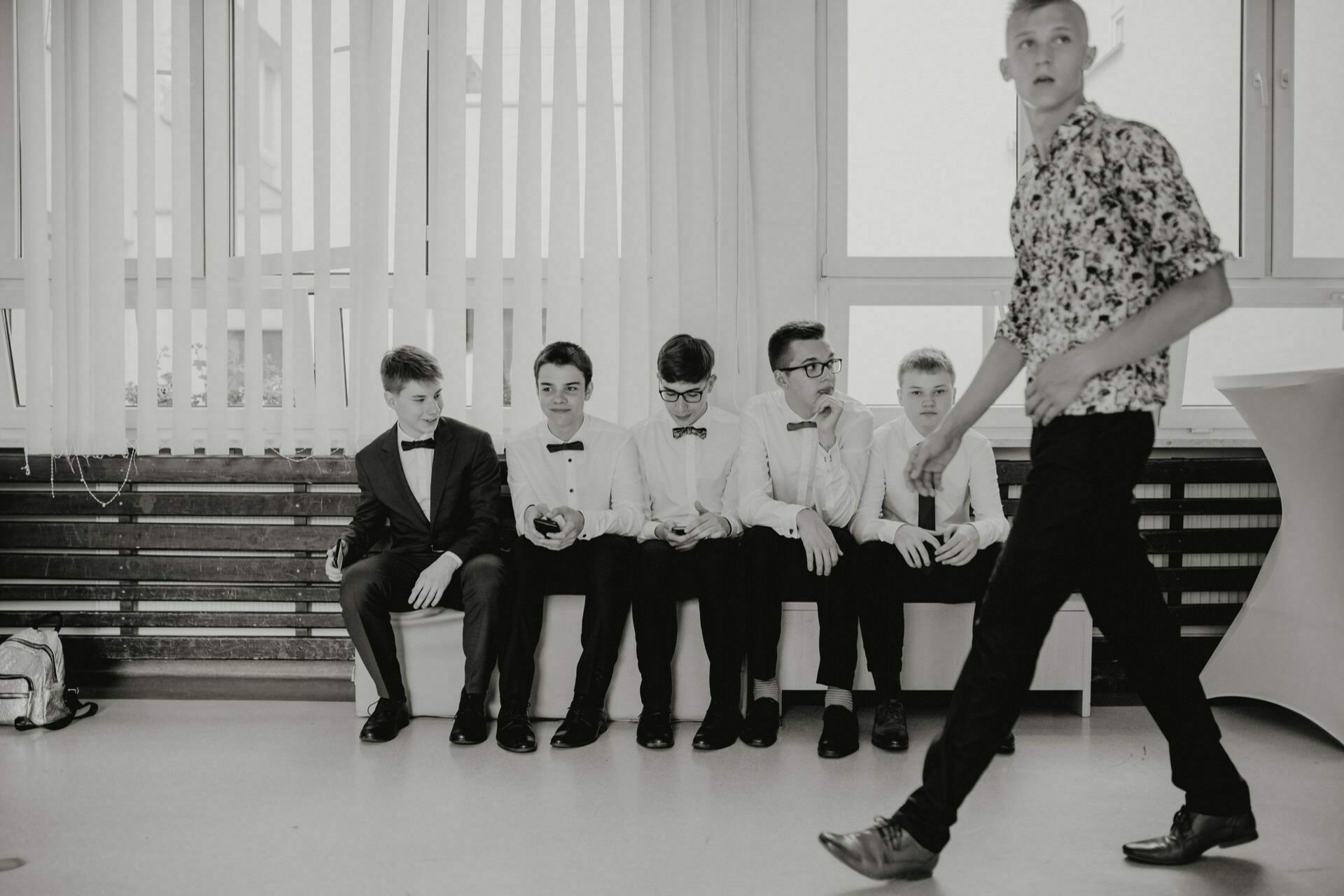 The black and white photo shows six young men, five of whom are sitting on a bench dressed in formal attire, with bow ties and suits. A sixth person walks by in the foreground, dressed in a floral shirt and dark pants. The venue appears to be an event taking place indoors - a perfect shot for event photography warsaw.  