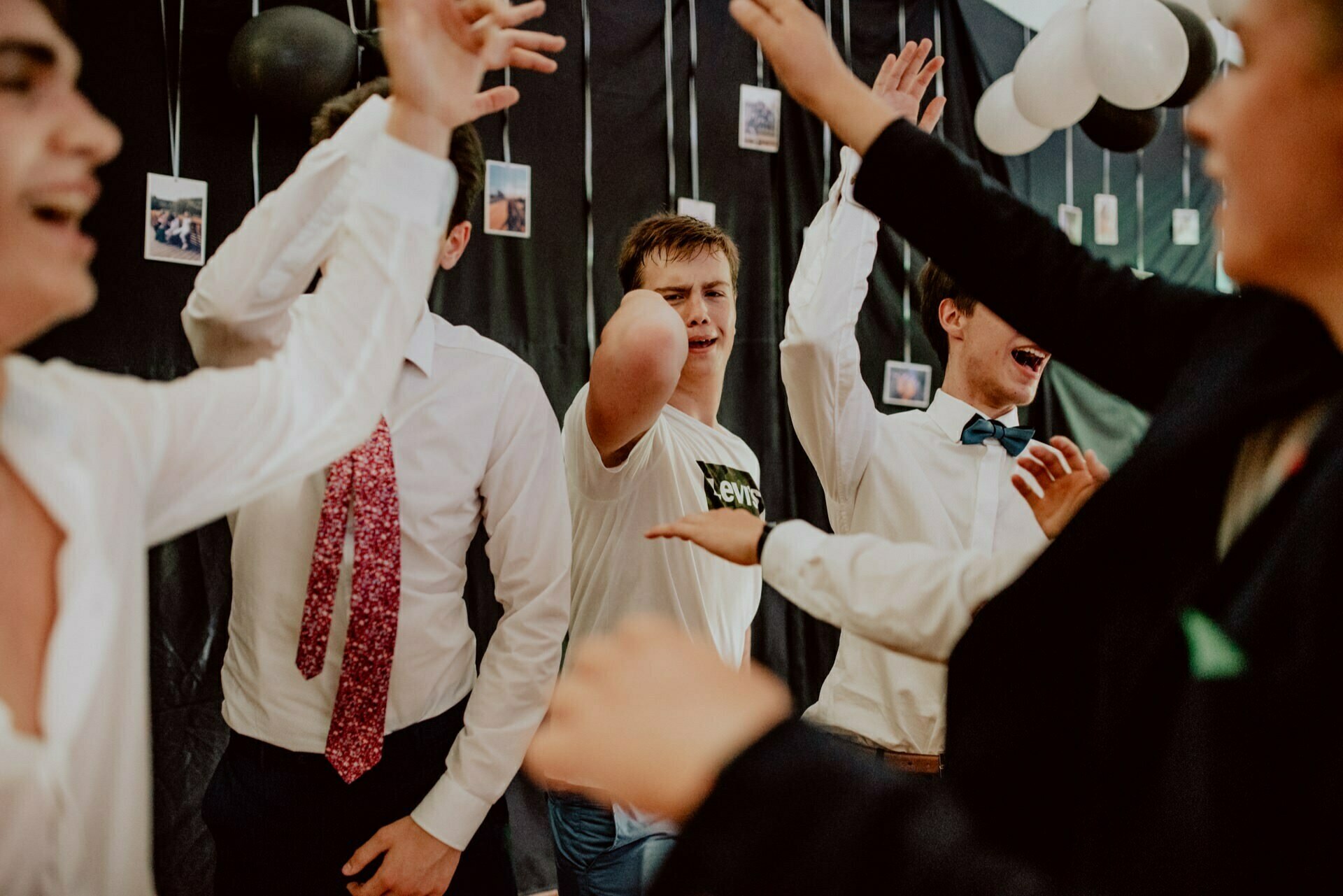 A group of people, some dressed in white shirts and patterned ties, enthusiastically dance and raise their hands in the air. The background is decorated with black fabric, black and white balloons and small photos hanging on strings. The mood is joyful and lively - perfect for capturing event photography.  