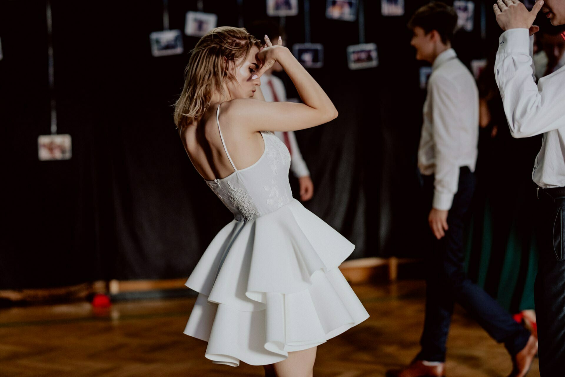 A woman in a white layered dress dances with her arms raised at a social event. Other people are also dancing, including a man in a white shirt and dark pants. A dark curtain can be seen in the background, with photos of the event's photoshoot hanging on it.  