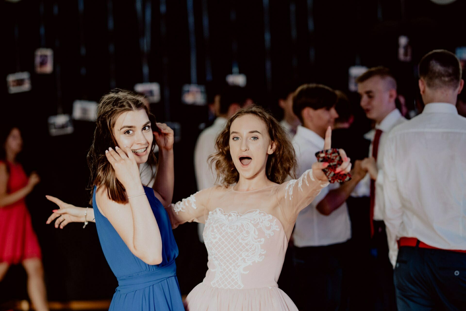 Two young women, one in a blue dress and the other in a light pink dress, assume playful poses at a dance party. Guests in formal attire mingle and dance under the low lighting and dangling photo decorations, perfectly capturing the essence of the photo event. 