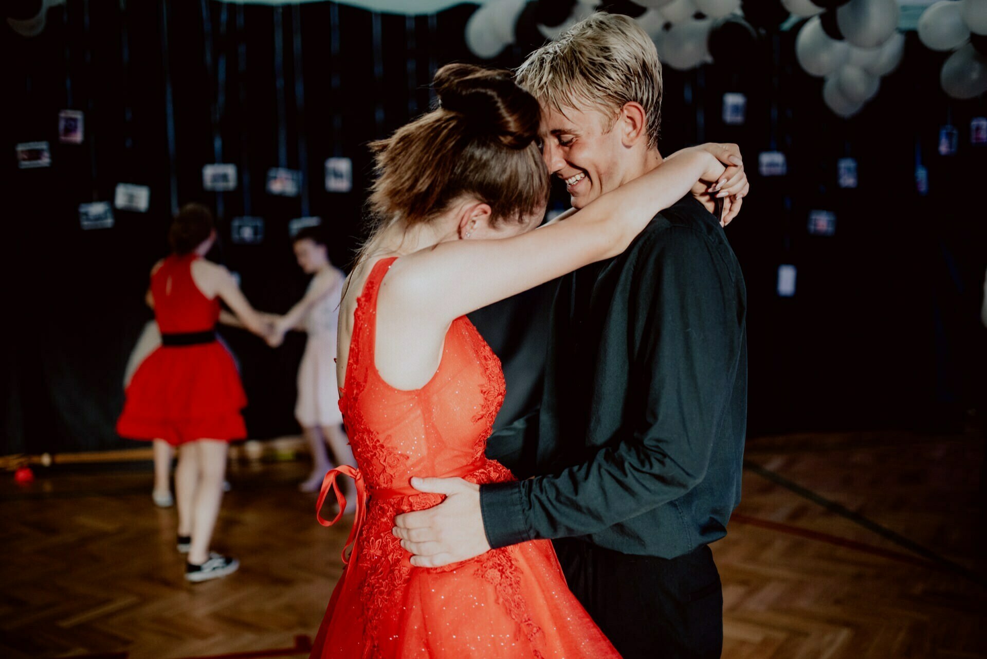 A young couple in formal attire, a woman in a red dress and a man in a black shirt, share a close dance in a dimly lit room decorated with balloons and photos. Other people dance in the background, beautifully captured by an event photographer Warsaw for an unforgettable photo report of the event. 