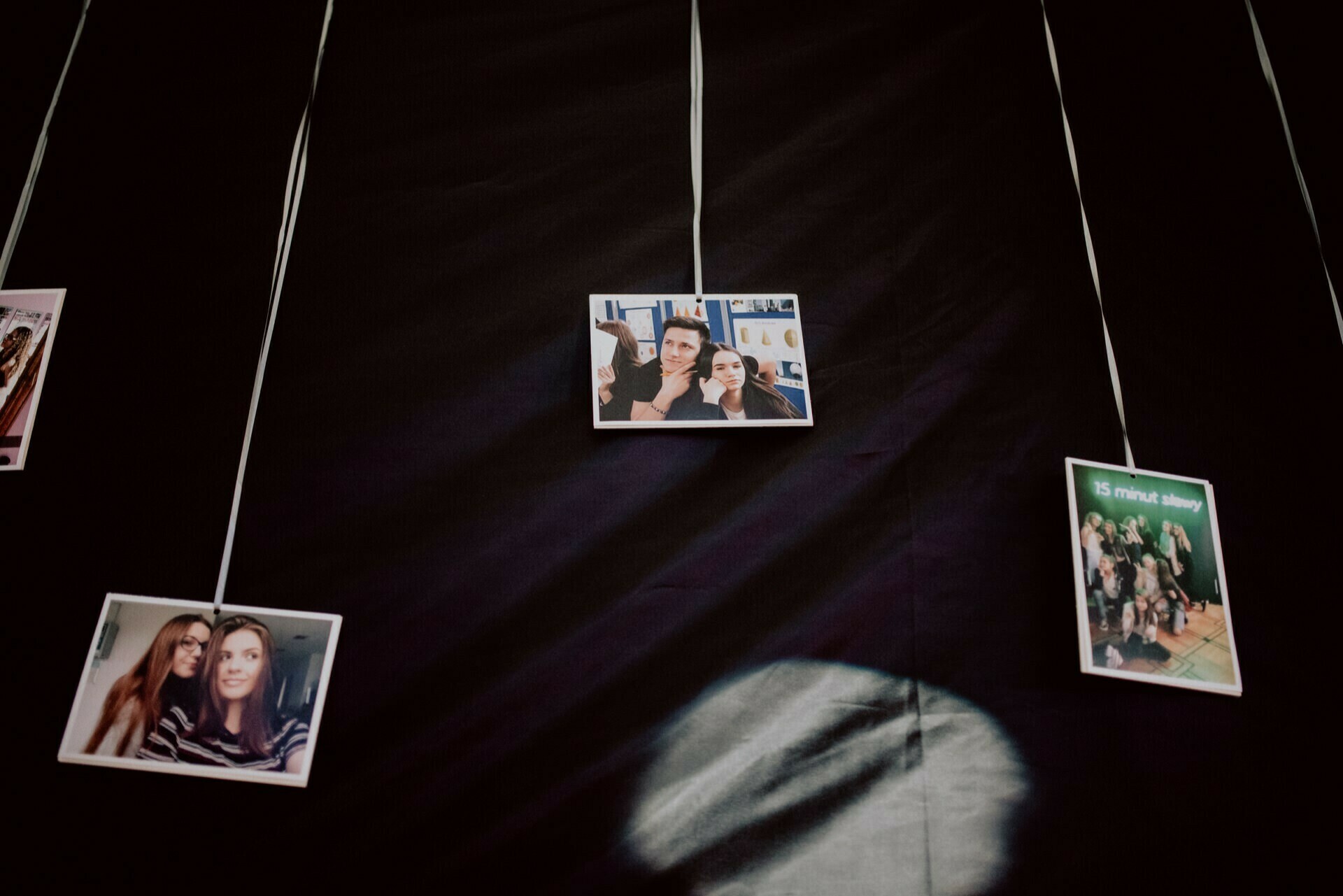 Three photographs hang against a dark background, suspended by white strings. The central photo shows two people posing for a selfie. The photo on the left also shows two people posing. The right photo, with a green title, is part of a photo report of the events and shows a group of people in a candid moment.   