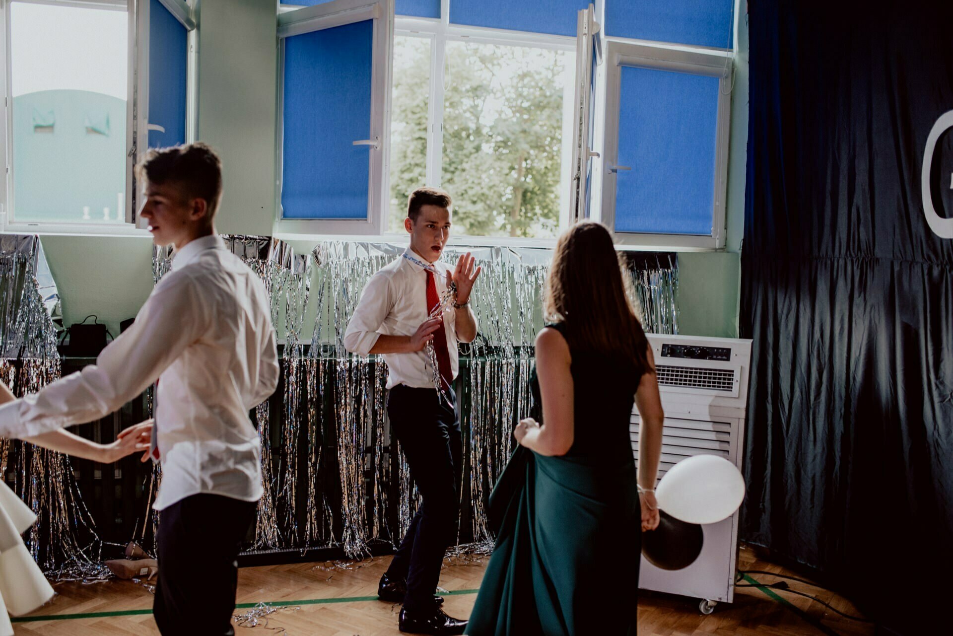 People in semi-formal attire dance in a decorated room with glittering curtains and large windows. One person in a white shirt and tie stands in the middle while others move around her. The atmosphere suggests a festive event, perhaps a dance or party - perfect for event photography.  