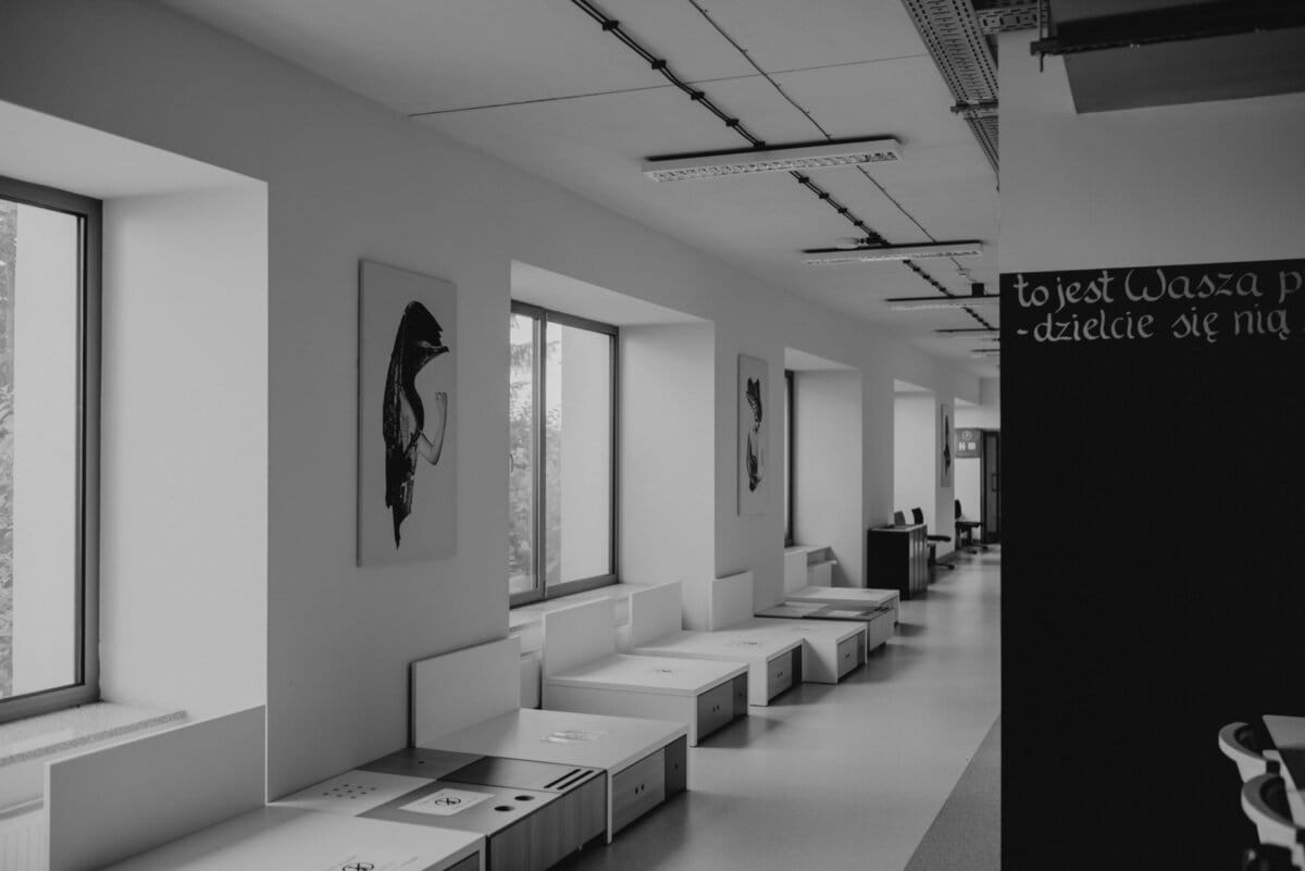 The black-and-white photo by Marcin Krokowski shows a modern corridor with large windows on the left and a wall of blackboard on the right. Under the windows are benches with built-in storage, decorated with works of abstract art hanging above them. 