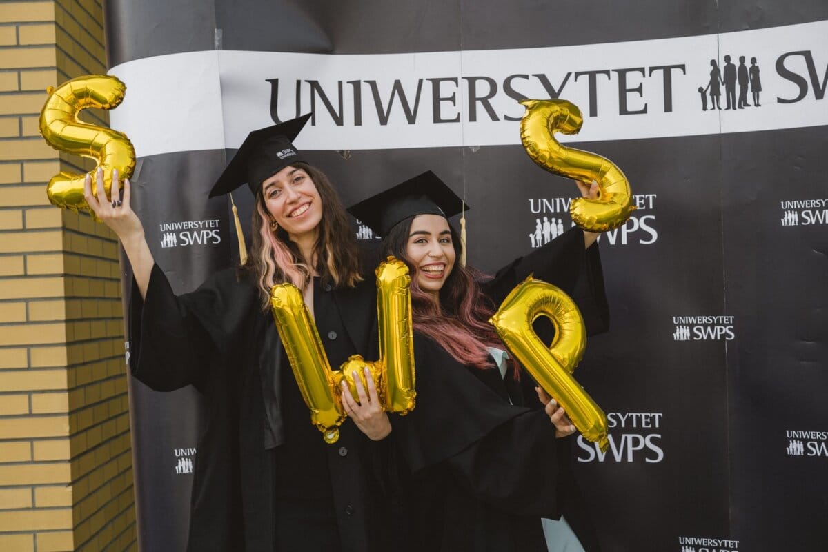 Two graduates in black caps and aprons stand in front of the "SWPS UNIVERSITY" sign. They hold large gold balloons forming "SWP," smiling and celebrating their achievement. This moment was brilliantly captured by Marcin Krokowski, an acclaimed event photographer Warsaw.  