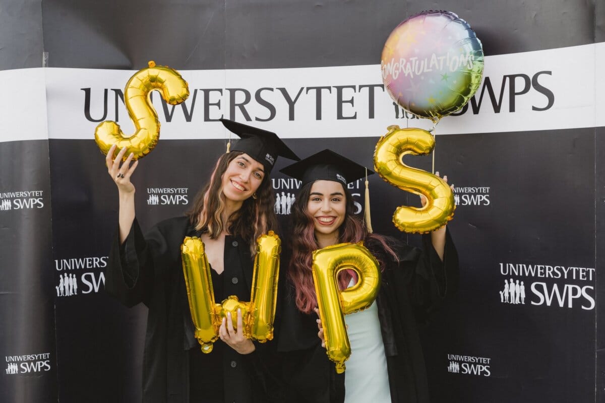 Two graduates wearing black caps and aprons are holding gold balloons with "SWPS" written on them against a background of "SWPS University." One of them is also holding a congratulatory balloon. Both are smiling radiantly, which was perfectly captured in a photo report of the events by Marcin Krokowski.  