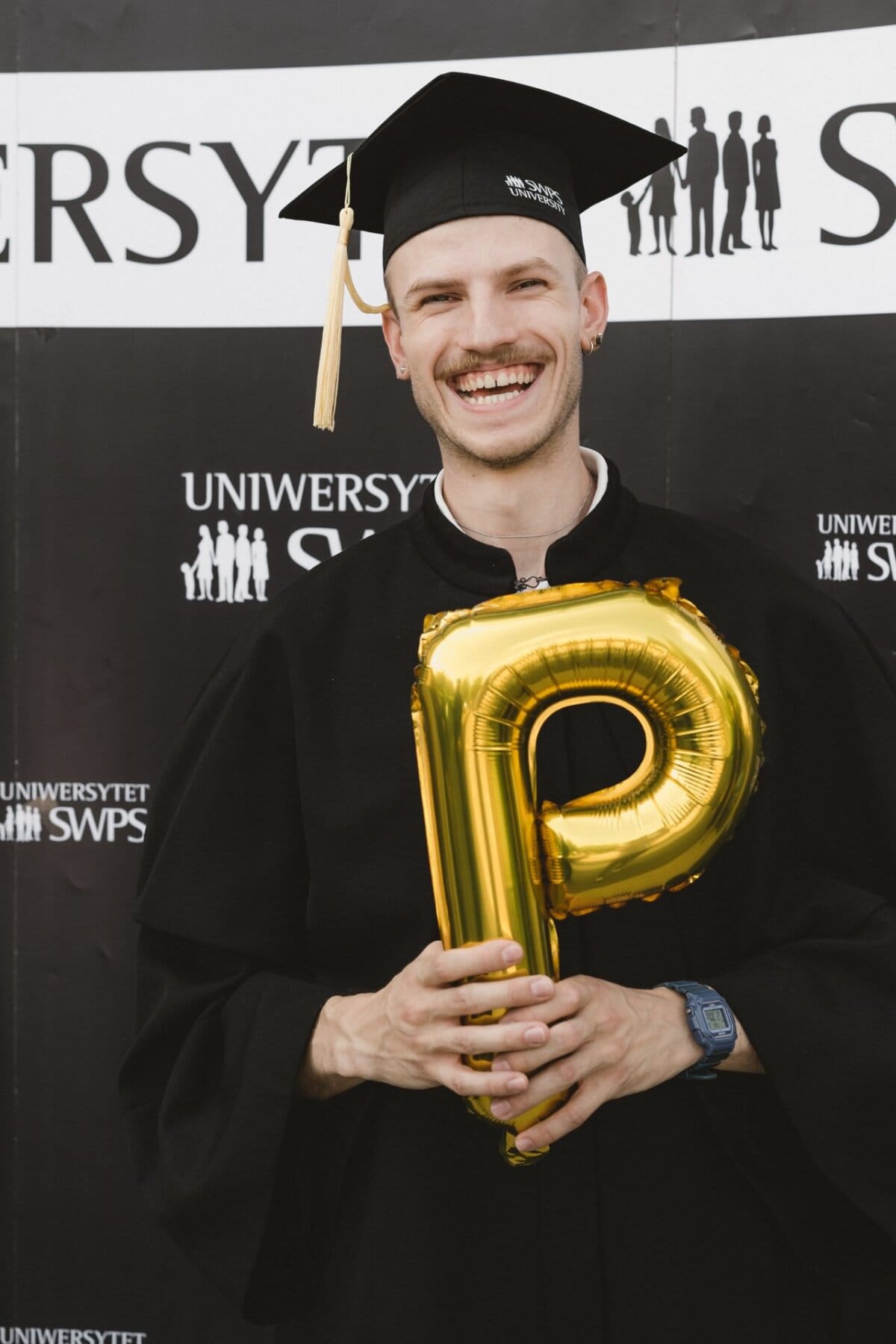 Wearing a graduate's outfit, including a black cap and toga, the person holds a gold balloon in the shape of the letter "P." He is standing in front of a banner with the words "SWPS UNIVERSITY" and a logo depicting silhouettes. The person is smiling widely, as if captured by Marcin Krokowski during an exciting photo shoot of the event.  