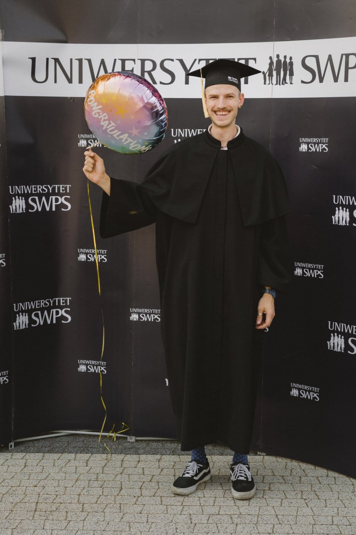 A person wearing a graduation cap and toga stands in front of a backdrop that reads "SWPS UNIVERSITY," holds a colorful balloon that reads "Congratulations!" and smiles. The image captures a joyful moment, fitting perfectly into any photo coverage of an event by a event photographer.