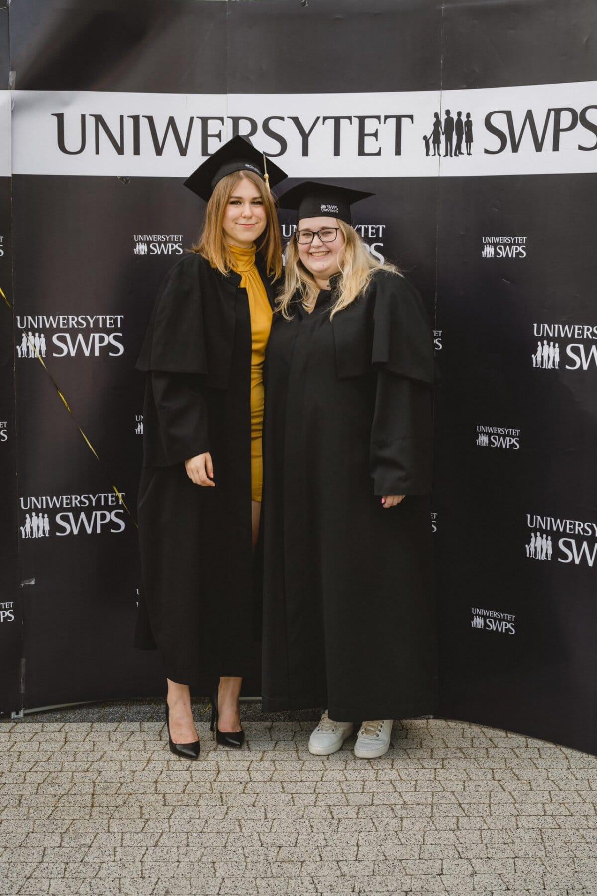 Two graduates in cap and toga stand together smiling at the repeated "SWPS UNIVERSITY." One is wearing a yellow dress and stilettos; the other is wearing a black outfit and white tennis shoes. The location seems reminiscent of an outdoor graduation ceremony - the perfect moment for event photography.  