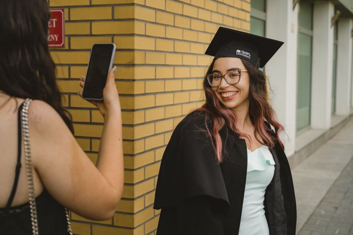 A graduate in a black cap and toga smiles as she poses for a photo taken by another person. The photo was taken outside against a yellow brick wall, capturing this special moment for the event photo essay. She holds the phone while another person preserves this precious memory.  