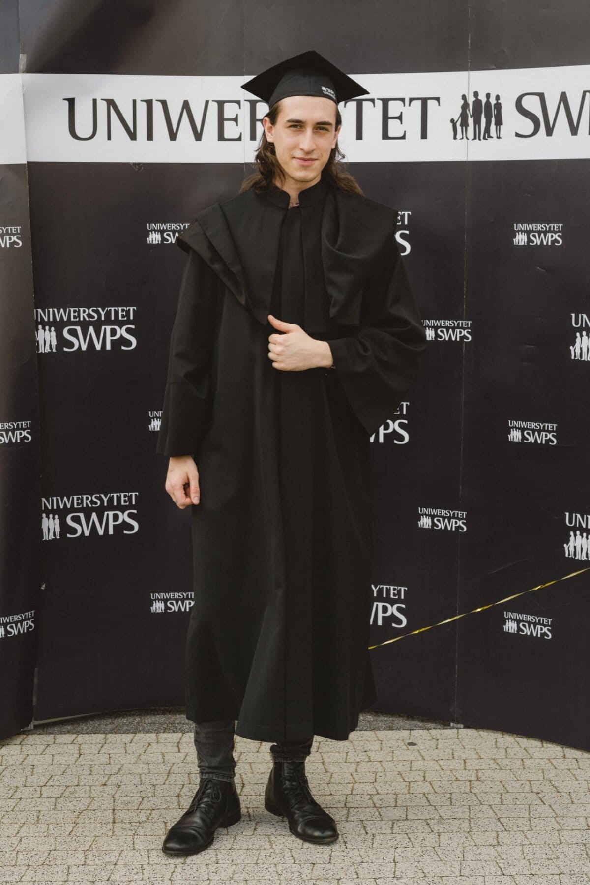 A graduate stands against a background decorated with "SWPS University" written repeatedly. Dressed in the traditional black cap and gown, they pose on the bright cobblestone tiles. This photo is part of an event photo essay that captures moments of the ceremony.  