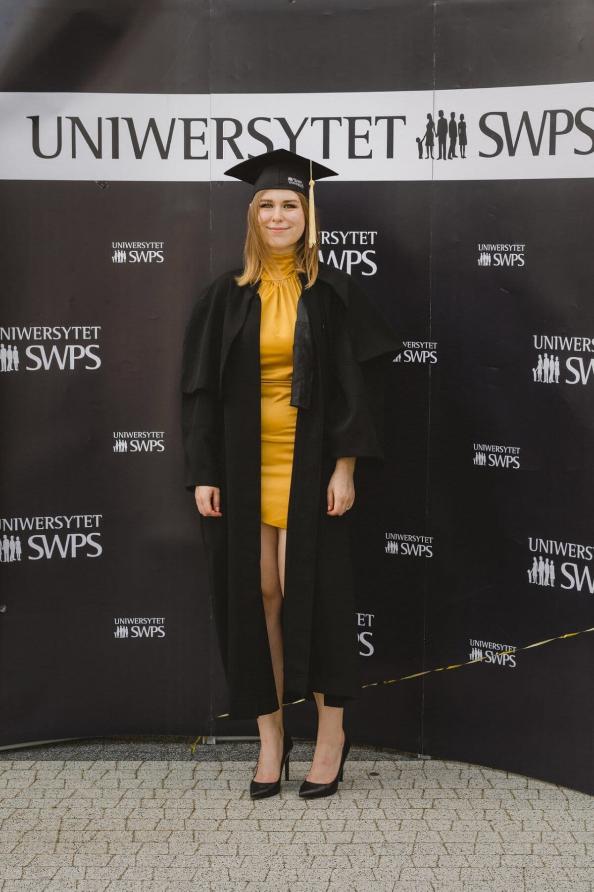 A young woman in a yellow dress and black bathrobe stands in front of a backdrop with the words "SWPS University." Captured by Marcin Krokowski, an event photographer Warsaw, wearing a black graduation cap and heeled shoes, she smiles at the camera. 