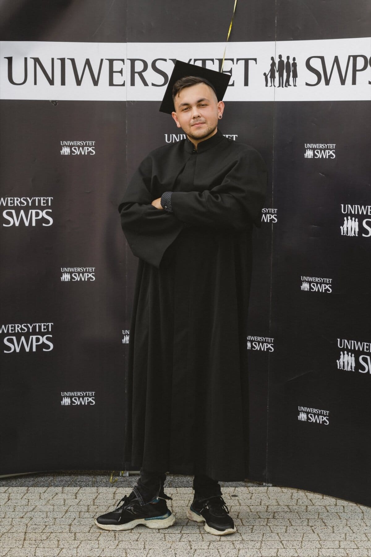 The person is standing against a background of "SWPS UNIVERSITY" logos, with his arms crossed, wearing a black cap and graduation gown, looking towards the camera. The background is black and depicts the graduation theme, capturing a memorable photo-report of the event. 