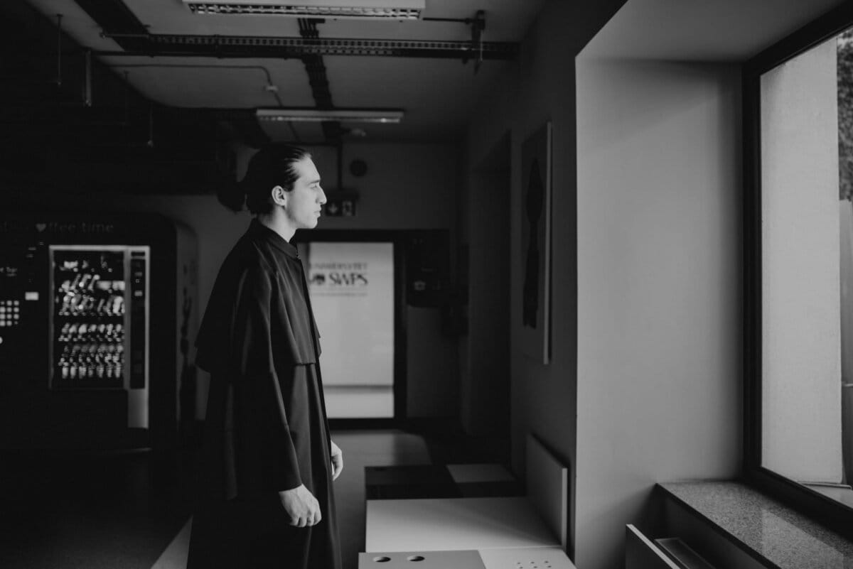 A person with long hair tied back stands in the room and looks out the window. They are dressed in dark, airy clothing. The scene is captured in black and white, creating a contemplative mood. An interesting element of the event photo report by Marcin Krokowski is the vending machine in the background.   