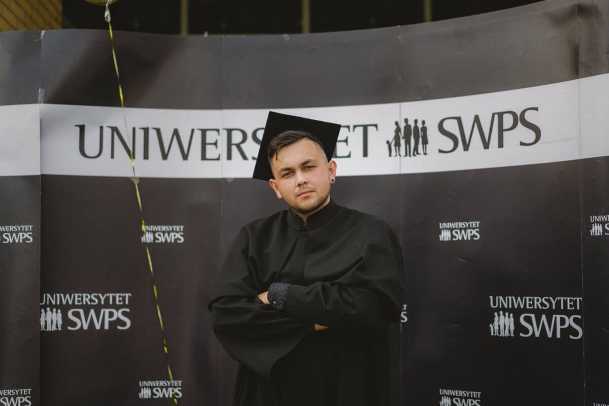 A person wearing a black graduation gown and cap stands in front of a background with the words "SWPS University." They are looking into the camera with their arms crossed. The photo, which is part of a photo report of the events by Marcin Krokowski, also shows a string of balloons in the upper left corner.  