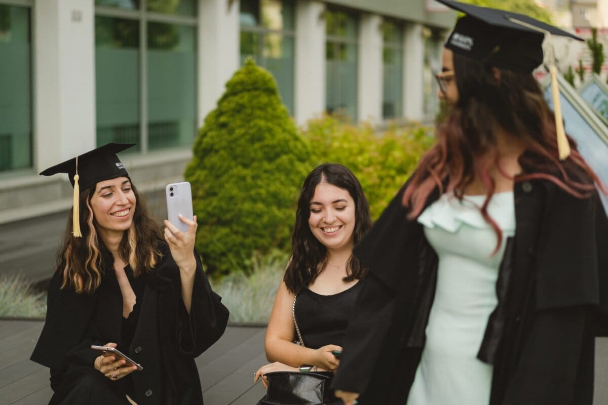 Three women are celebrating graduation. One sits wearing a graduation cap and holding a smartphone, smiling at another woman standing in cap and gown. The third woman sits in the middle and smiles without her cap. They are outside, surrounded by greenery, beautifully captured in Marcin Krokowski's event photography.   