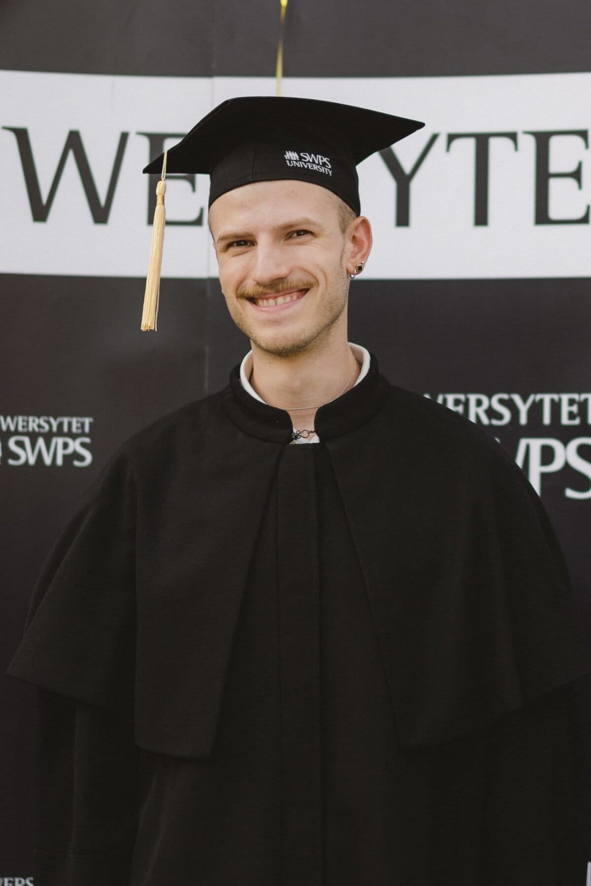 A person smiles at the camera, dressed in a black graduation gown and a cap with a yellow clutch. They are standing against a background of fragmentary text "SWPS UNIVERSITY." They have short hair, a bright mustache, earrings and a tie under their gown - an example of professional event photography.  