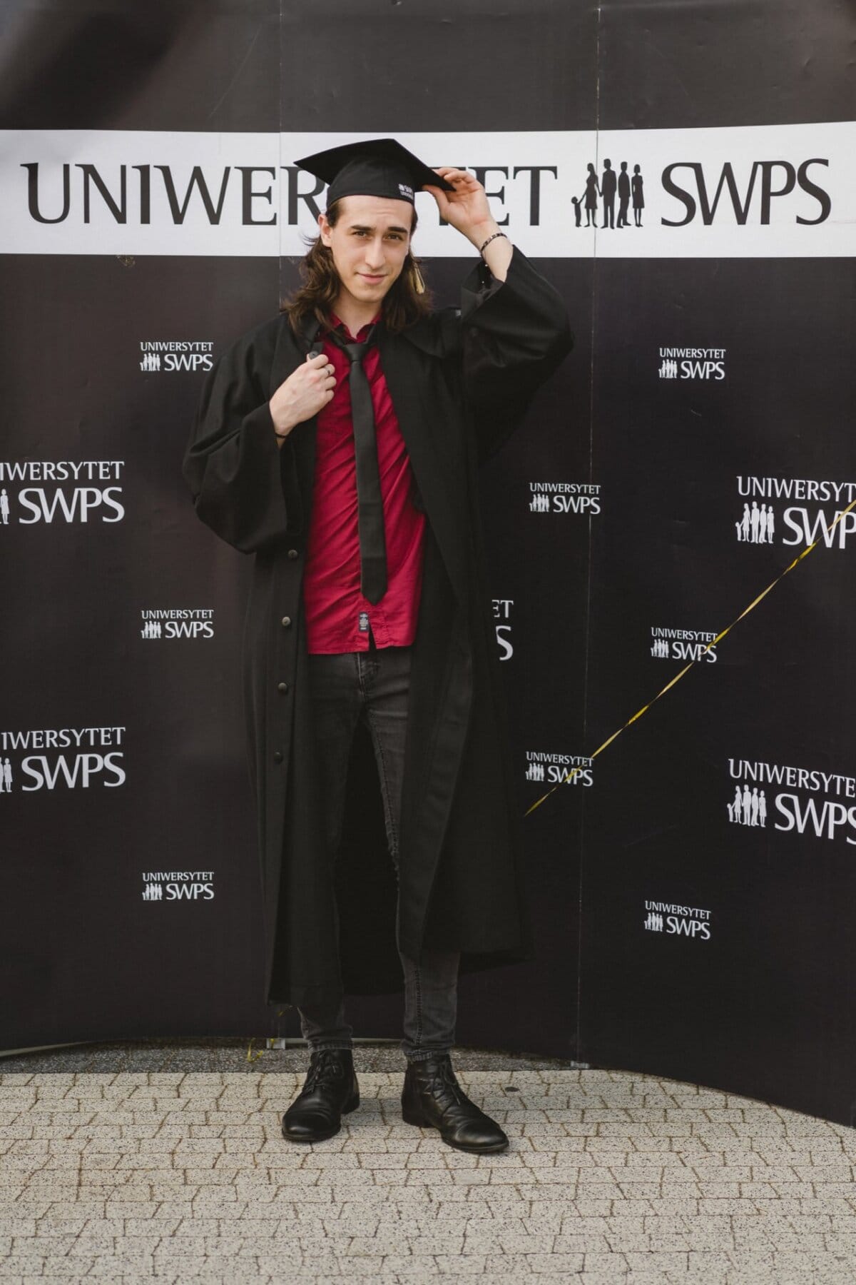 A young person with long hair poses at a graduation ceremony wearing a black dress and cap. They are standing against a background with the SWPS University logo. The person captured by event photographer Marcin Krokowski is wearing a red shirt, black tie and black shoes.  