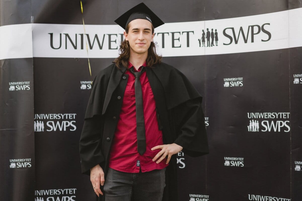 A person with long hair, wearing a graduation cap and gown imposed over a red shirt, poses confidently with his hand on his hip. In the background is a "SWPS UNIVERSITY" banner with the university's logo and a small balloon in the upper left corner. Marcin Krokowski captures this memorable moment in his photo report of the events.  