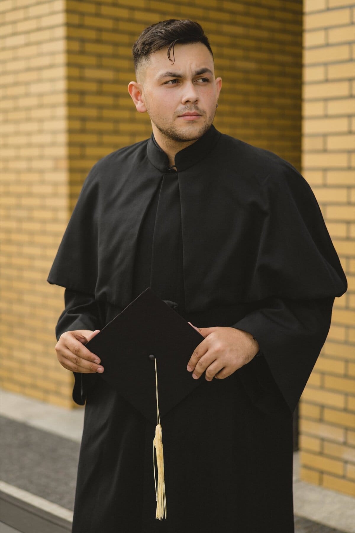 A young man in a black graduation gown holds a cap with a pom-pom. He stands in front of a brick wall and looks into the distance with a serious expression, looking at the seemingly sunny day. This event photography perfectly captures the importance of the moment.  