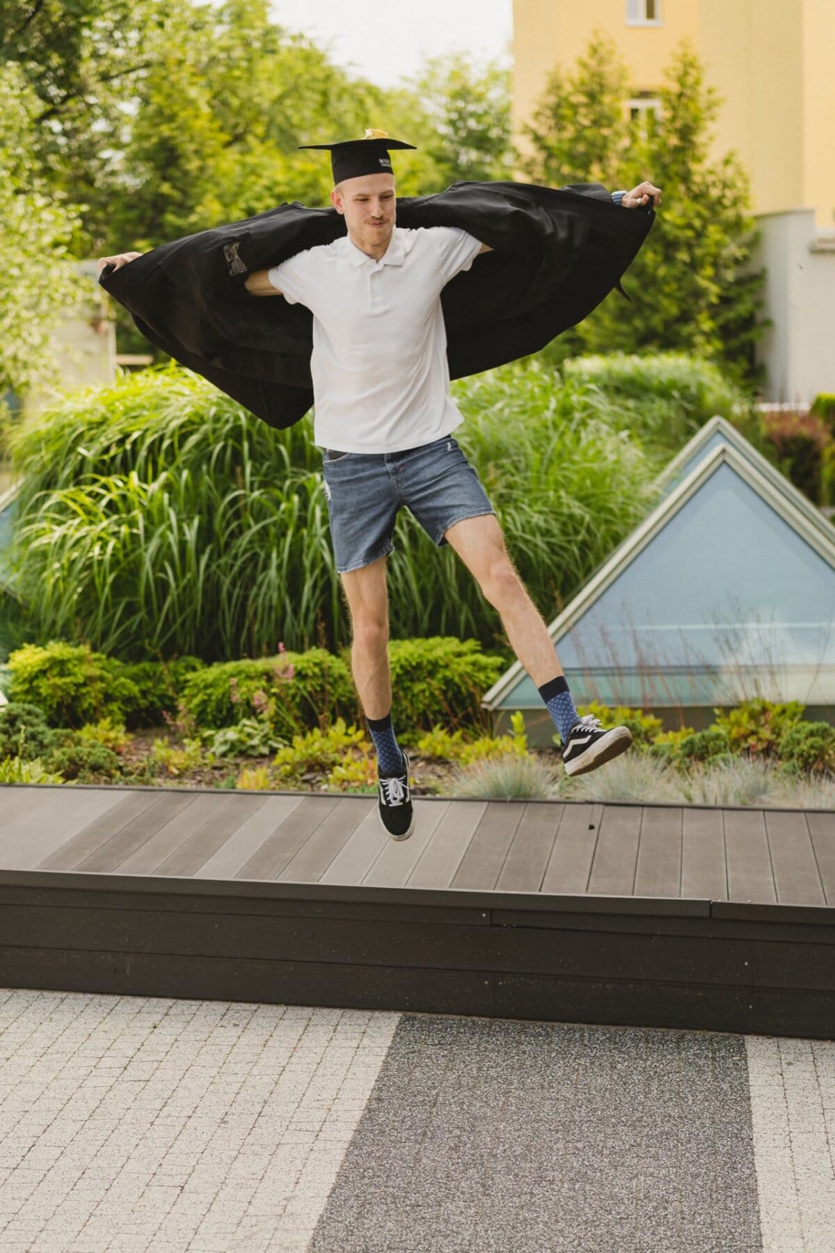 A young man wearing a graduation cap, polo shirt, shorts and tennis shoes joyfully jumps in the air outside. His arms are outstretched, holding his jacket wide open behind him, against a backdrop of greenery and modern rooftops. The scene captures the essence of Marcin Krokowski's event photojournalism.  