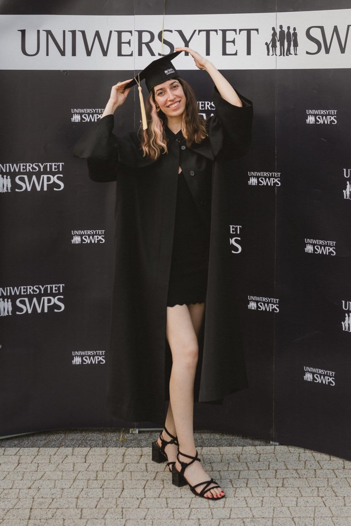 A graduate in a black graduation gown and cap stands in front of "SWPS University." With one hand, she holds the tassel of her cap and smiles. She is also dressed in a black dress and stiletto heels, with one leg slightly bent, as part of a photo recap of the events.  
