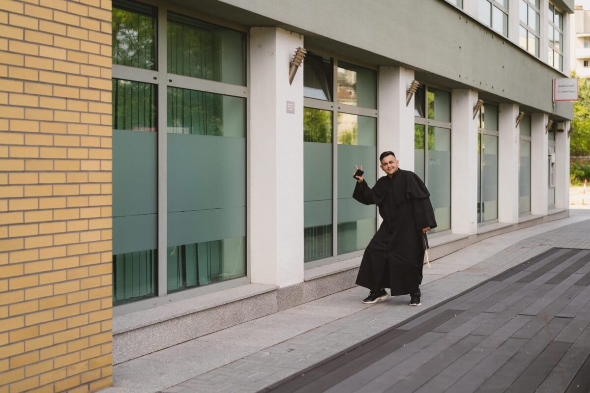 A person wearing a long black coat and sneakers smiles and makes a peace sign with his hand, standing on the sidewalk in front of a building with large windows. This event photo captures the moment perfectly, with the person's bag in the other hand and the brick wall to the left. 