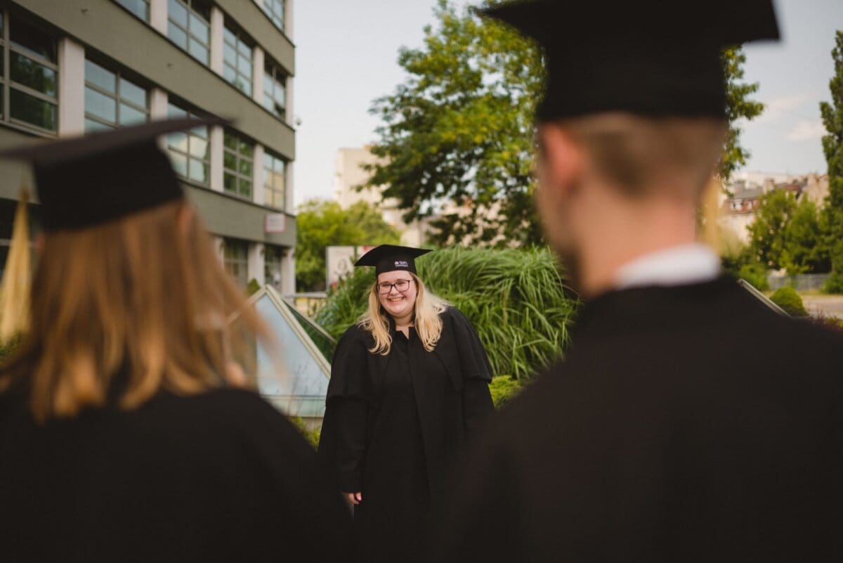 A person wearing a cap and graduation gown stands smiling outside, facing two other people dressed in similar graduation attire, with his back to the camera. The building and lush greenery can be seen in the background, capturing a memorable photo of the event by Marcin Krokowski. 