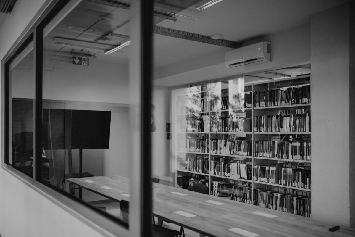 A black and white photo of a conference room with a long table, several chairs and a large TV screen. Next to the room is a library with shelves filled with books. The modern space, captured by Marcin Krokowski, is well lit with windows and overhead lamps.  