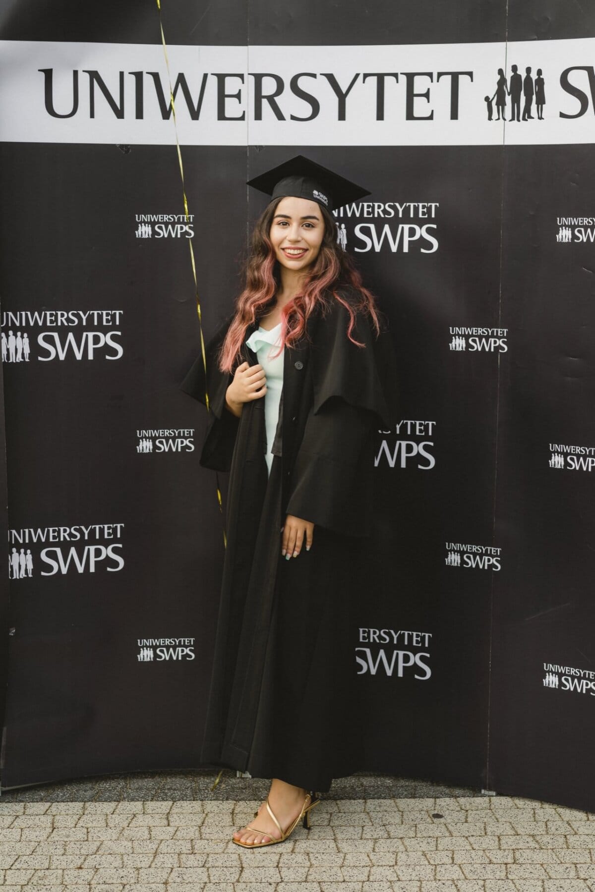 A woman wearing a cap and graduation gown stands against a black background with the words "SWPS UNIVERSITY" written several times. She has long, wavy hair and is smiling. She is holding a yellow balloon string and wearing sandals. This event photography beautifully captures the joy of the event.   