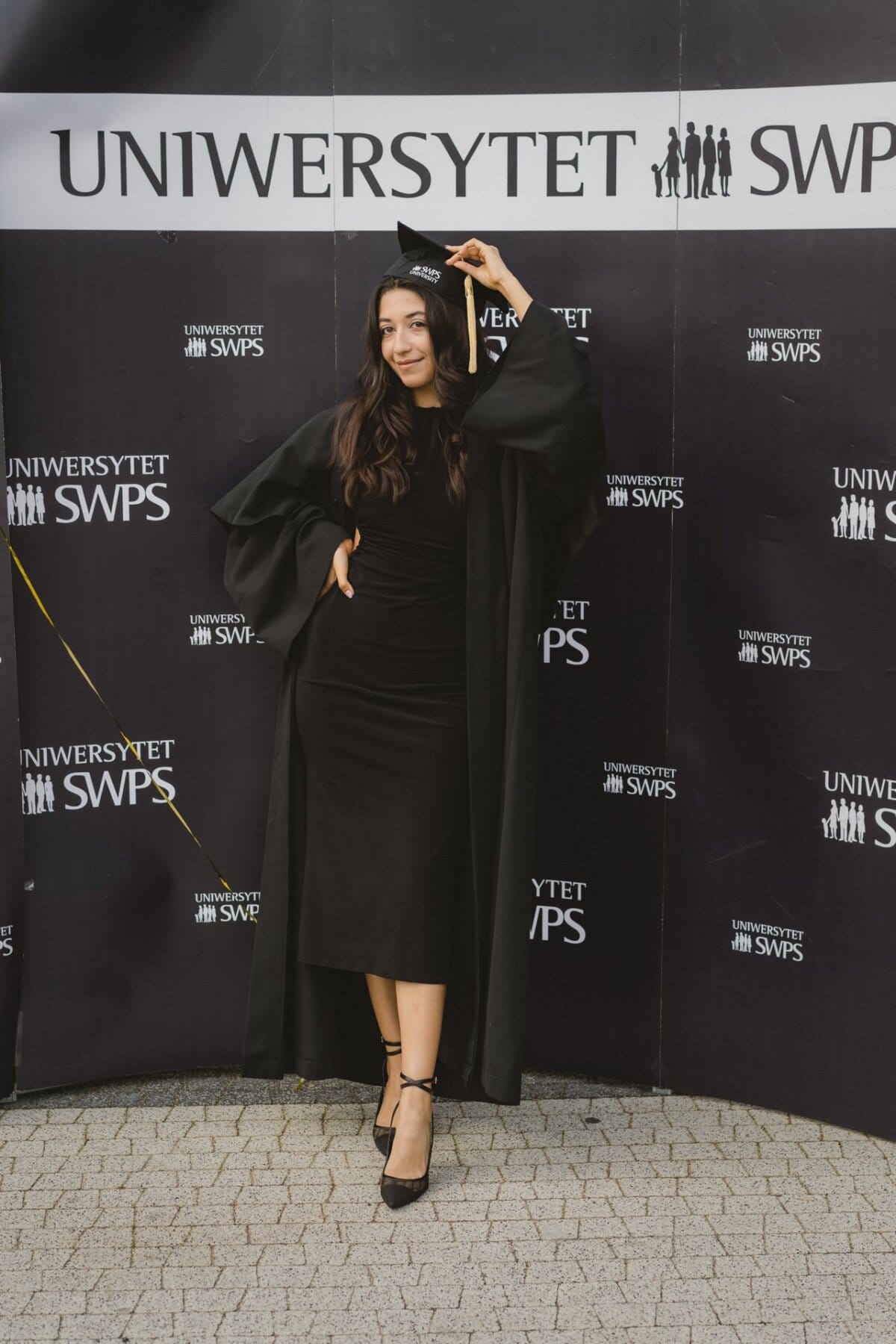 A graduate in a black cap and toga poses happily against the backdrop of the "SWPS UNIVERSITY" sign. She improves her cap with one hand, stands confidently on the paved surface, wearing black stilettos - capturing the moment brilliantly with event photography. 