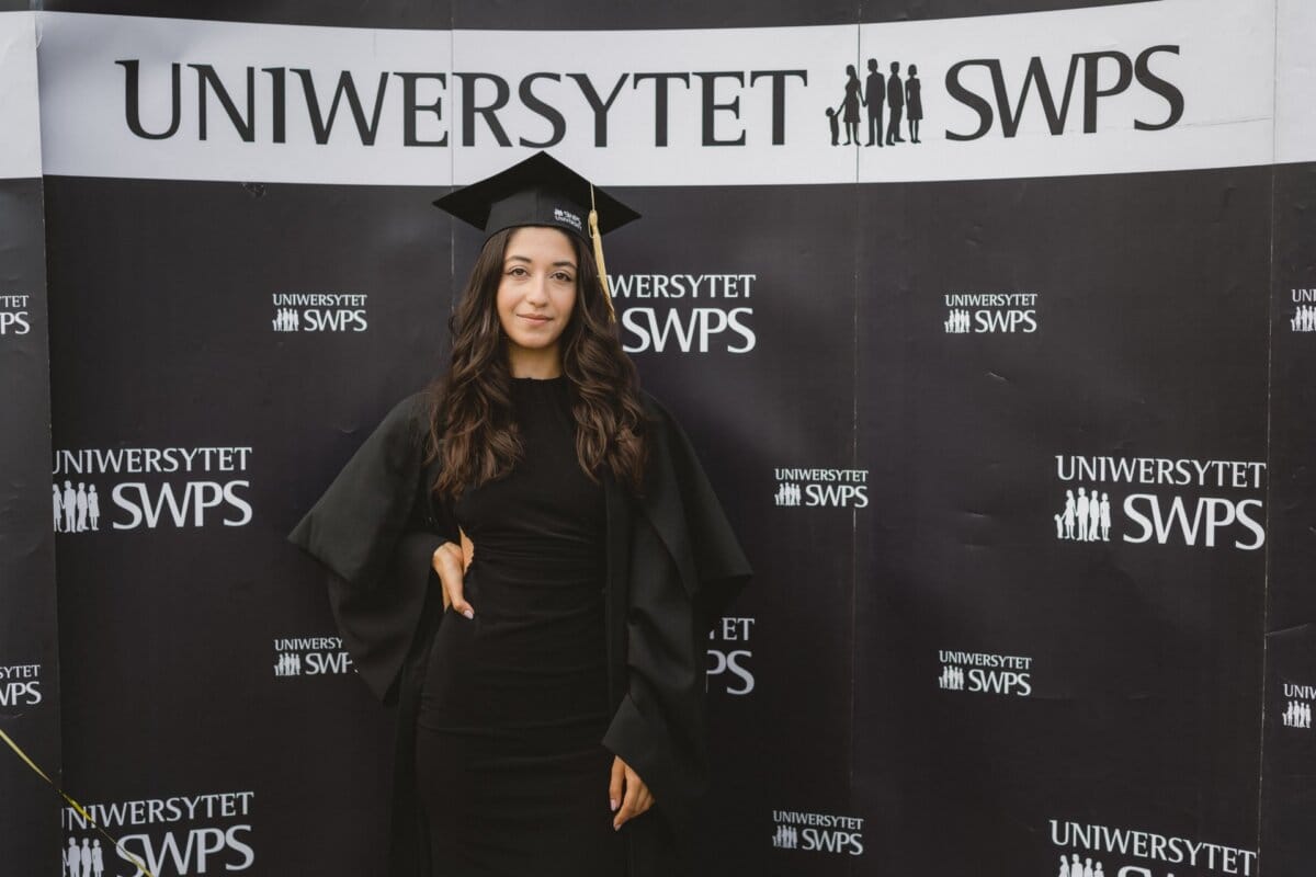 The graduate, wearing a black cap and toga, stands in front of a black backdrop that repeatedly reads "SWPS UNIVERSITY." He has long, dark hair and smiles at the camera. In the background are small white logos and text, perfectly captured by event photographer warszawa, Marcin Krokowski.  