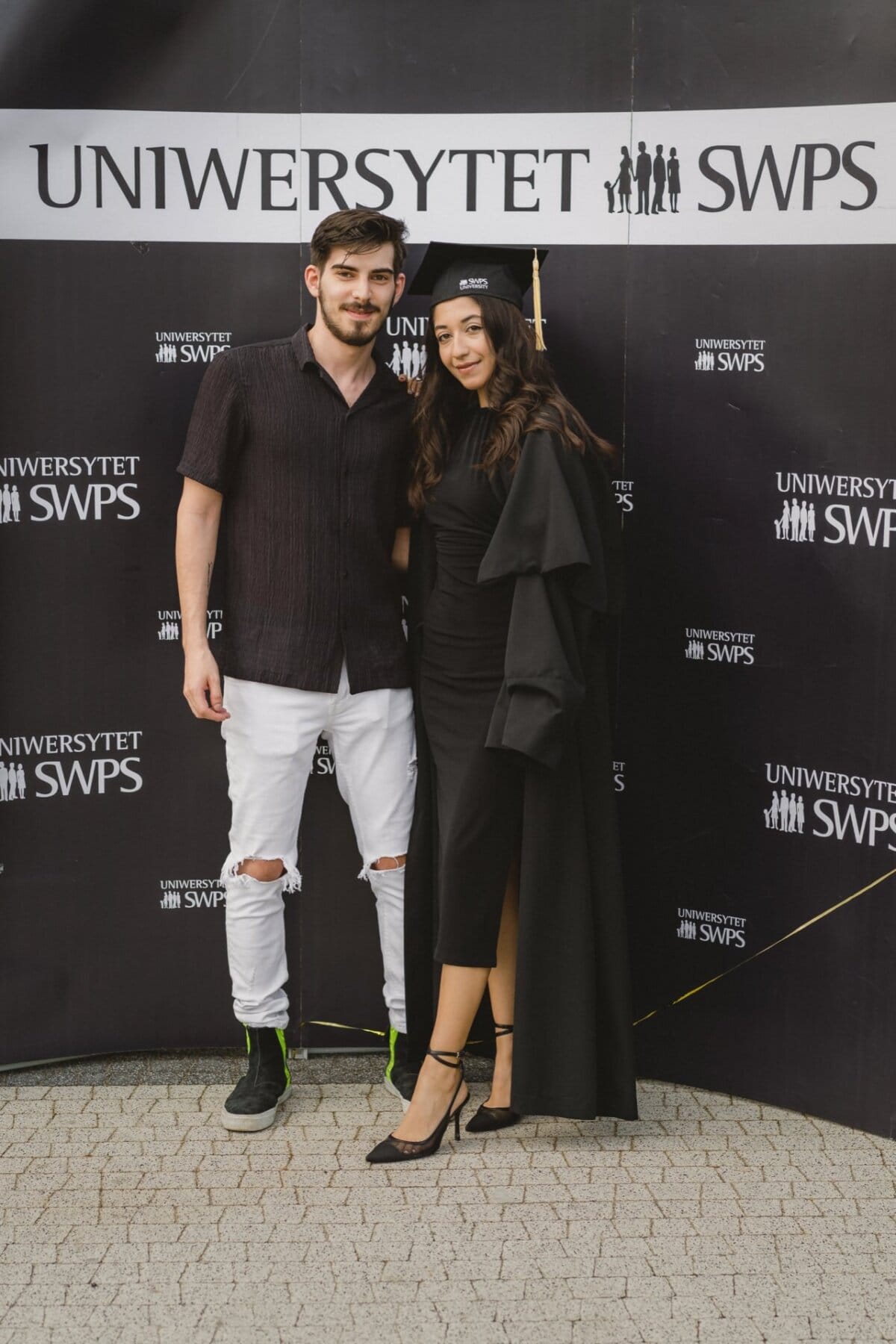 A man and a woman stand in the background of the SWPS UNIVERSITY. The woman is wearing a cap and graduation gown, the man is dressed in a black shirt and white ripped jeans. Both are smiling at the camera, capturing the moment in an event photo.  