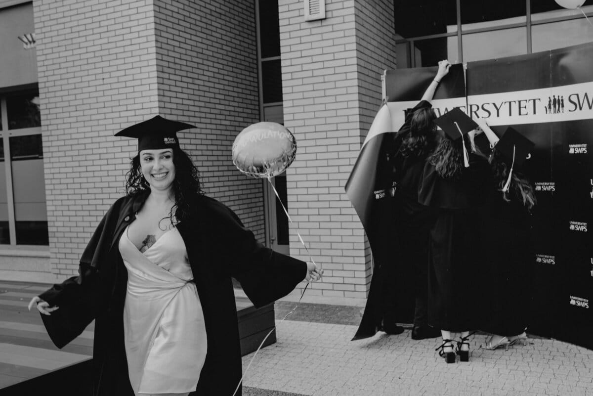 A graduate in cap and gown smiles while holding a balloon that reads "Congratulations." Behind her, a group of people in graduation attire sets up a backdrop with the words "SWT UNIVERSITY." The scene shows a lively photo recap of the event outside the brick building.  