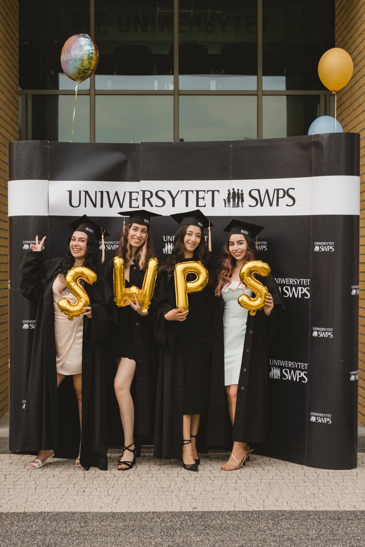 Four women in graduation gowns and caps stand in front of the "SWPS University" banner. They hold gold balloons forming the letters "SWPS." One wears a hat with balloons attached, and another makes the peace sign. Everyone is smiling and celebrating their achievements in this vibrant photo essay of the event.   