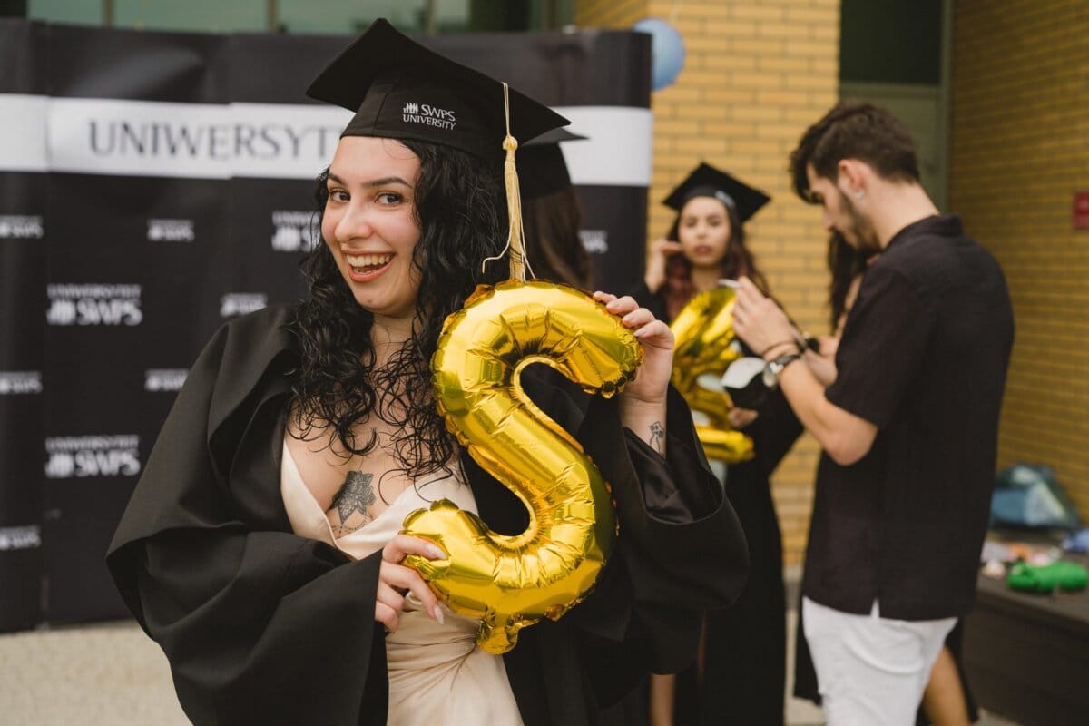 A graduate in cap and gown holds a gold balloon in the shape of the letter "S". He stands in front of a partially visible "University" sign. Other graduates in caps and gowns are celebrating in the background, capturing the moment as part of a photo recap of the events.  