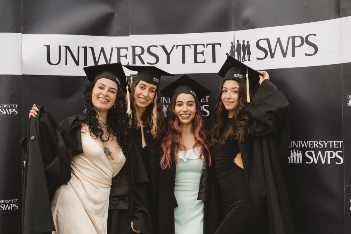 Four people wearing caps and aprons are smiling and posing together in front of the "SWPS UNIVERSITY" sign. They look happy and celebratory, probably having just graduated. It's a perfect photo-op of the events.  