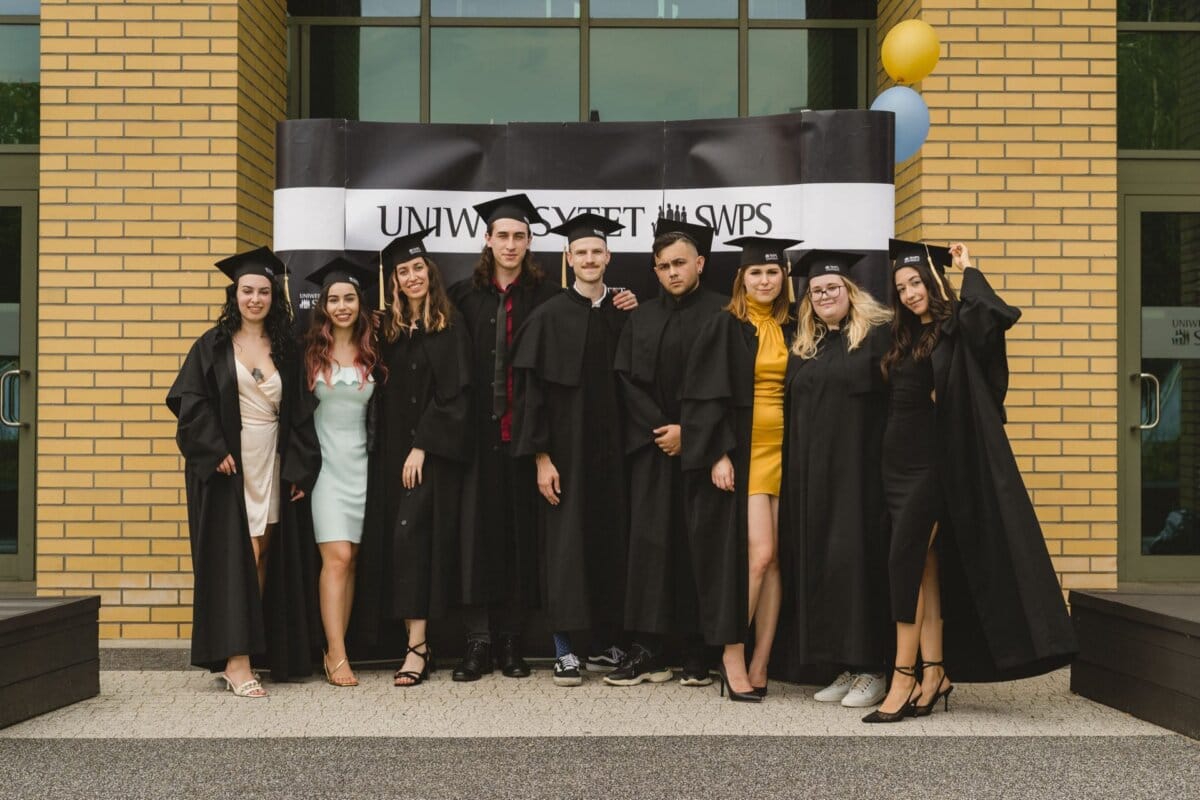 A group of nine graduates dressed in black academic togas and caps stand in front of the building with a banner reading "SWPS UNIVERSITY." They smile and pose together, celebrating their achievement. A photo of the event captures two balloons attached to the right side of the banner.  