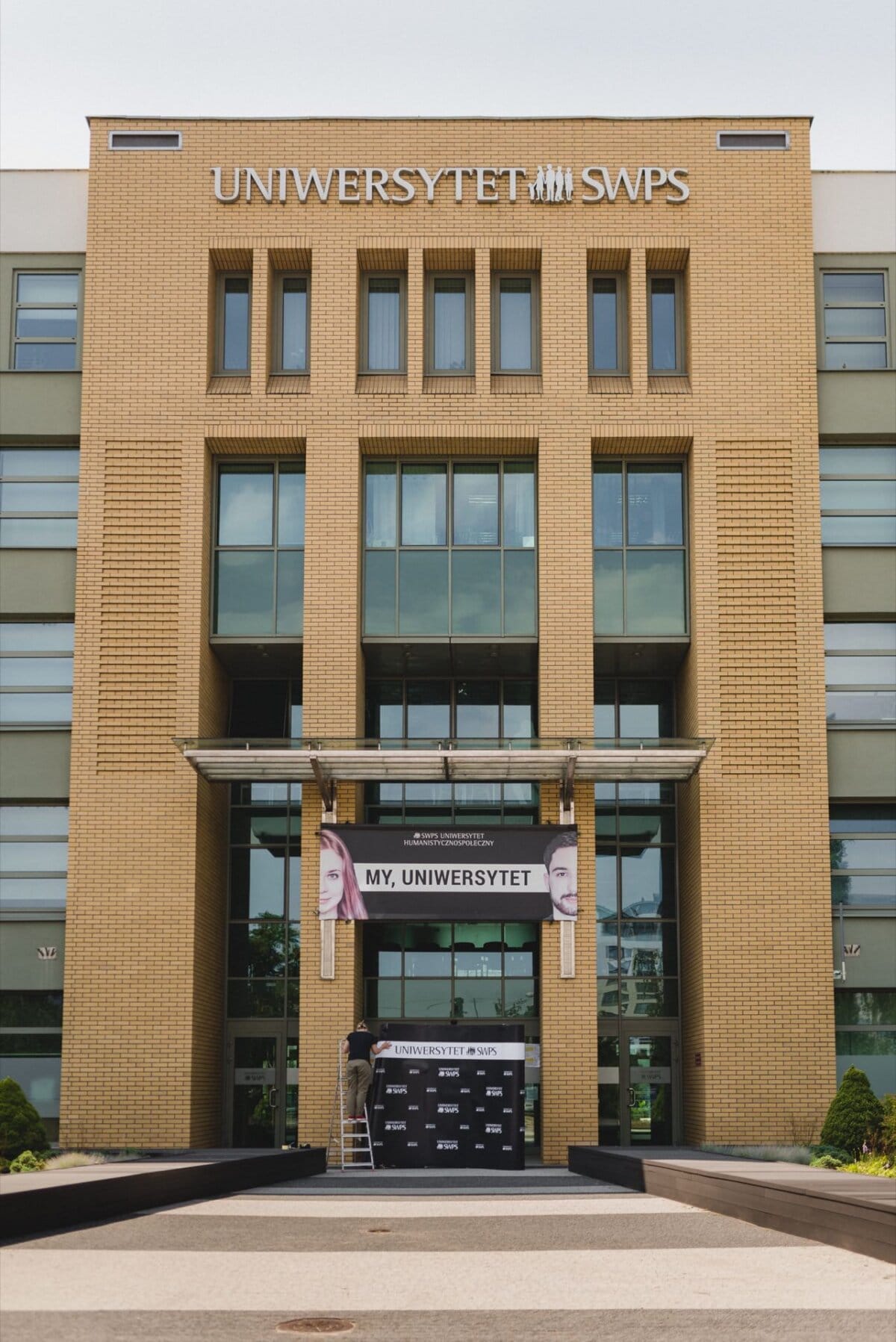 A person stands on a ladder and places a banner with the words "MY, UNIVERSITY" near the entrance to a modern, multi-story brick building with glass windows. Captured as part of a photo report of the events, the building bears the inscription "SWPS UNIVERSITY" on its facade. 