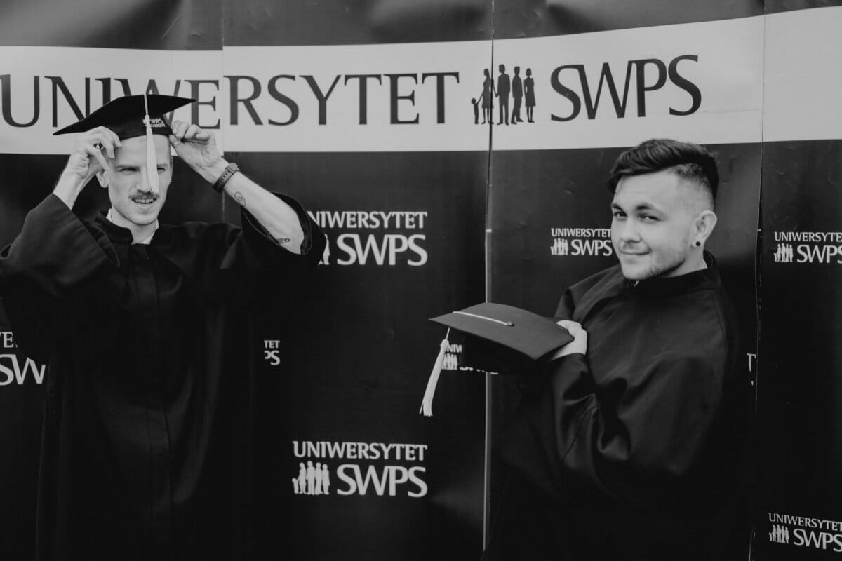 Two people wearing graduation caps and aprons against a background of the repeatedly printed "SWPS UNIVERSITY" sign. One person corrects the cap, while the other holds it and poses with a slight smile. This black-and-white photo captures a memorable moment from Marcin Krokowski's event photography.  