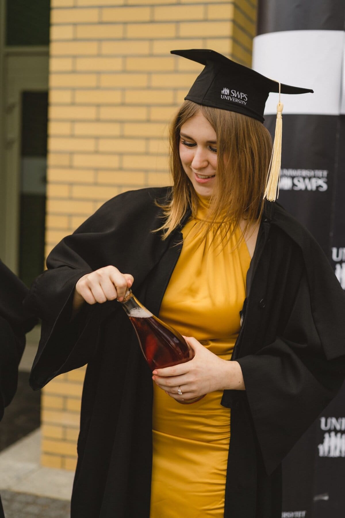 A person wearing a black gown and graduation cap holds and opens a bottle with a red liquid inside. The person stands in front of a brick wall and celebrates his achievement. This vivid moment captures the essence of Marcin Krokowski's event photography.  