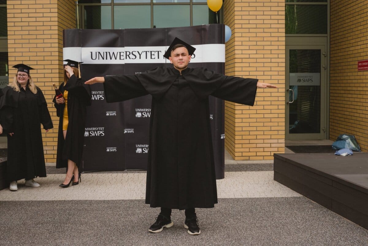 A person wearing a graduation cap and toga stands with his arms outstretched against the backdrop of the university, which reads "SWPS UNIVERSITY." Other graduates in caps and gowns can be seen in the background, beautifully captured by the event photographer. 