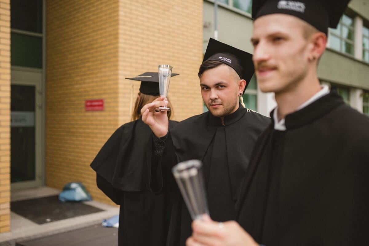 Three graduates in black gowns and mortarboards stand outside. One of the graduates in the middle faces the camera, holding a trophy-like object. The other graduates are partially visible, one also holding a similar object. In the background, a brick building captures an unforgettable moment from a photo recap of the events.   