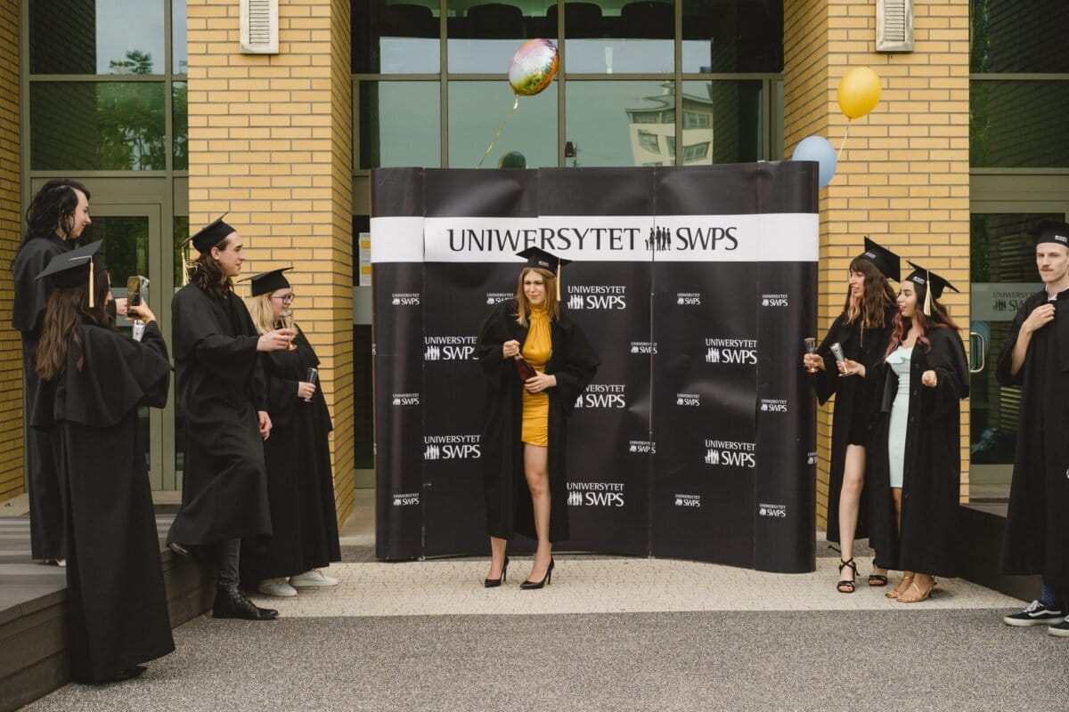 A group of graduates in black caps and aprons stand around a woman in a yellow dress and celebrate against the backdrop of the "SWPS UNIVERSITY" sign. Some are holding balloons, while others are gleefully gesturing, which was beautifully captured as part of the photo report of the events. 