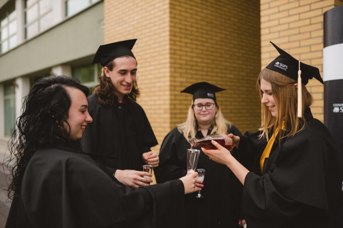 Four graduates dressed in black dresses and mortarboards celebrate together. Two women pour drinks into glasses while another woman and a man watch. They stand outside near the yellow brick-walled building and capture the joy of the moment in a beautiful photo report of the events by Marcin Krokowski.  