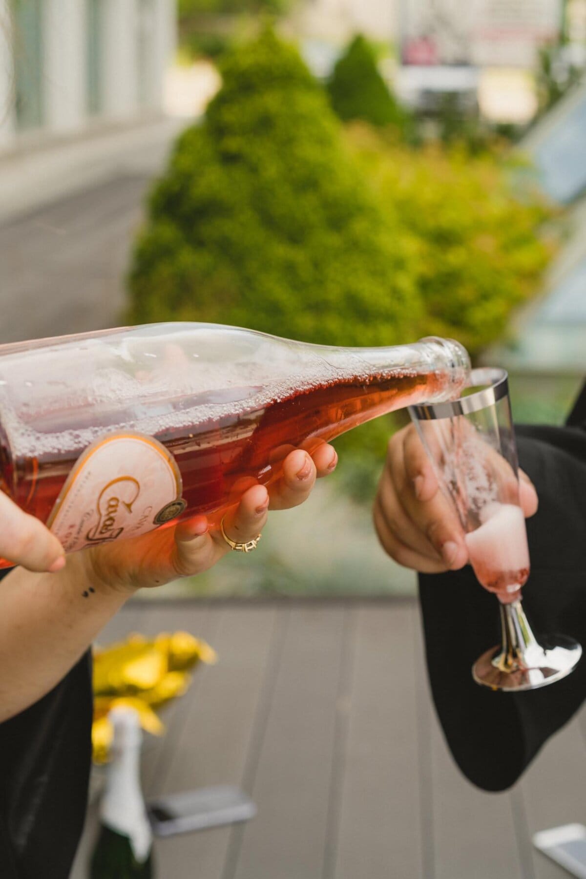 A person pours sparkling rosé wine from a large glass bottle into a fluted glass held by another person. Outside, there are green bushes and a building, creating a picturesque backdrop. The mood is festive, perfect for an event photo shoot.  