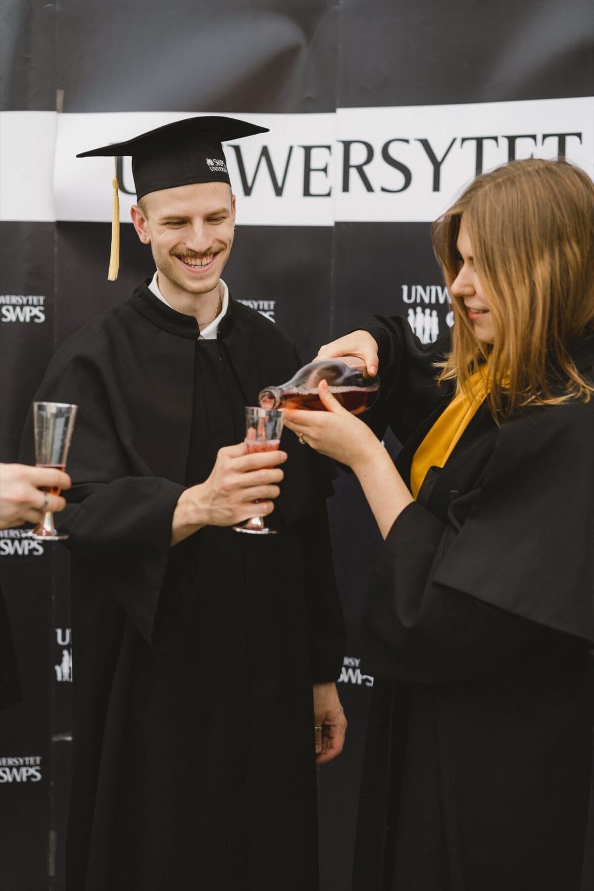 A smiling graduate wearing a cap and apron holds a glass while another graduate, also wearing a cap and apron, pours a drink into it. They are standing against a background of "UNIVERSITY" sign and other text. The hand of another person holding the glass is visible in the foreground, capturing an engaging photo essay of the events.  